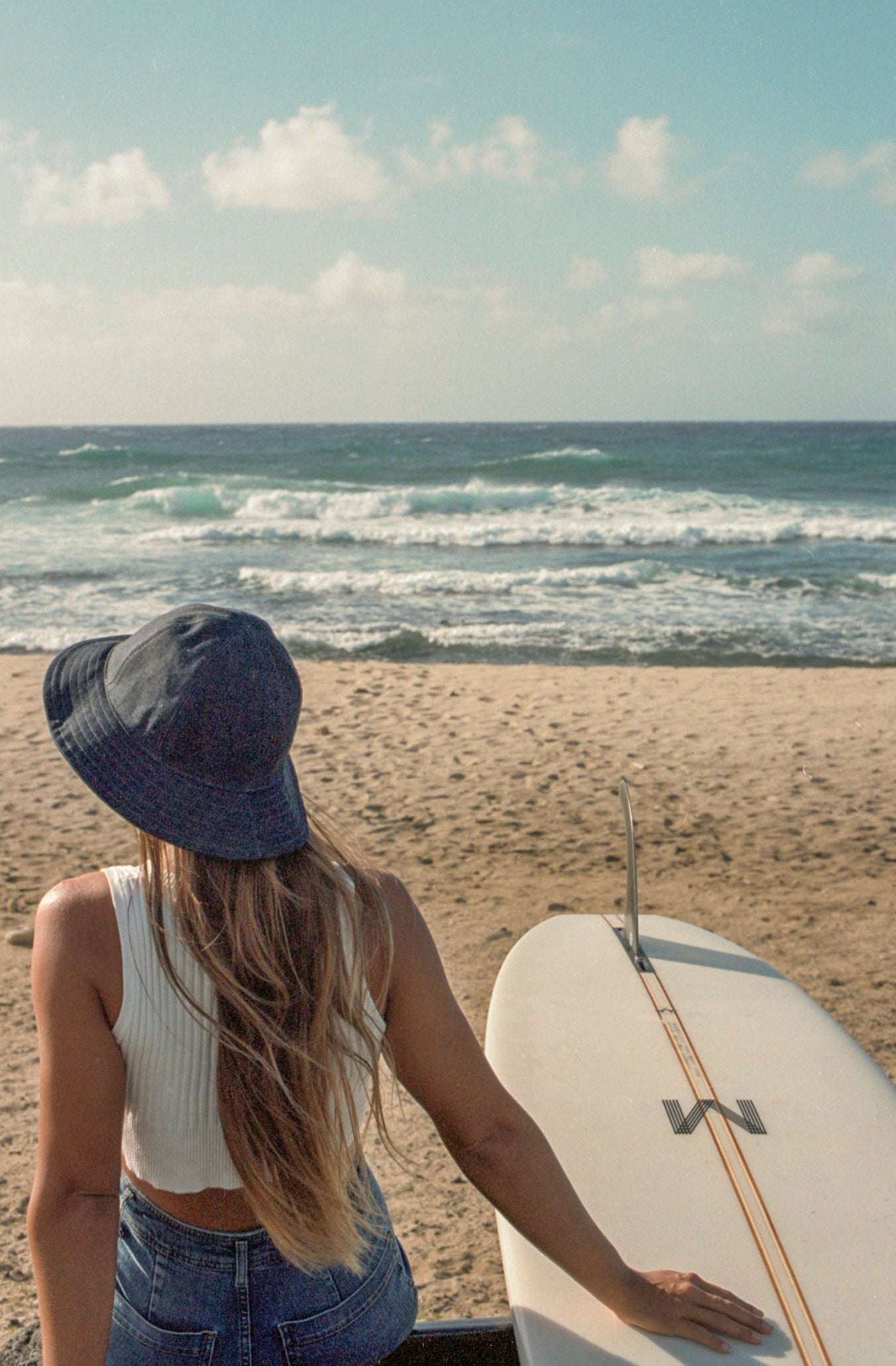 Girl wearing denim bucket hat with her surfboard, watching ocean waves.