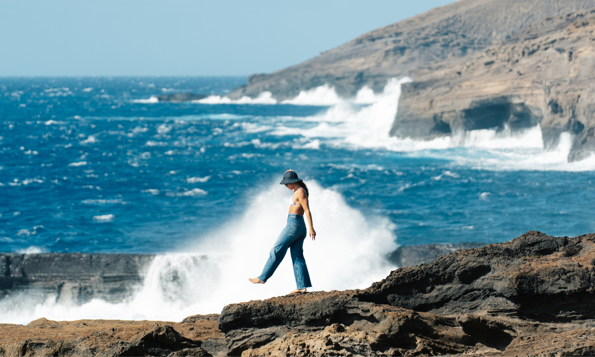 Girl wearing denim bucket hat walking on rocks, with an ocean view.